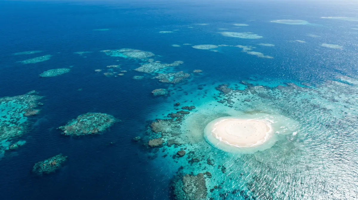 N aerial photograph of turquoise reef patches and a white sand cay scattered across deep blue water of the Great Barrier Reef off Queensland, bright midday light.