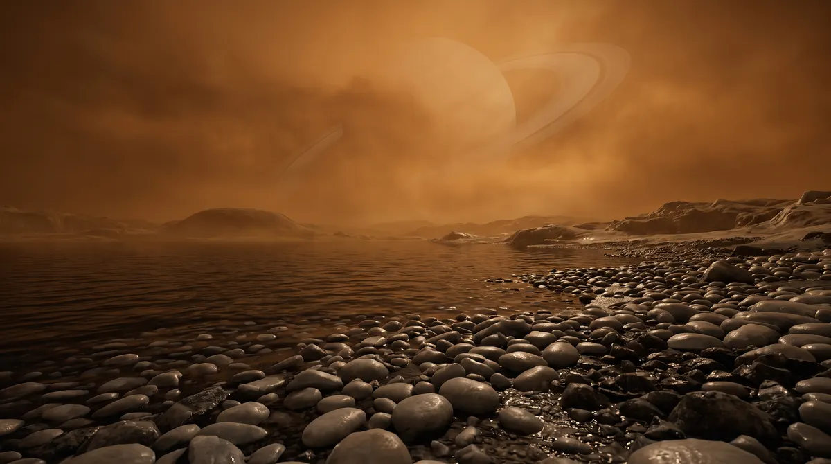 Landscape on Titan: a dark methane lake and rounded ice pebbles under a hazy orange sky, with Saturn faintly visible through the murk.
